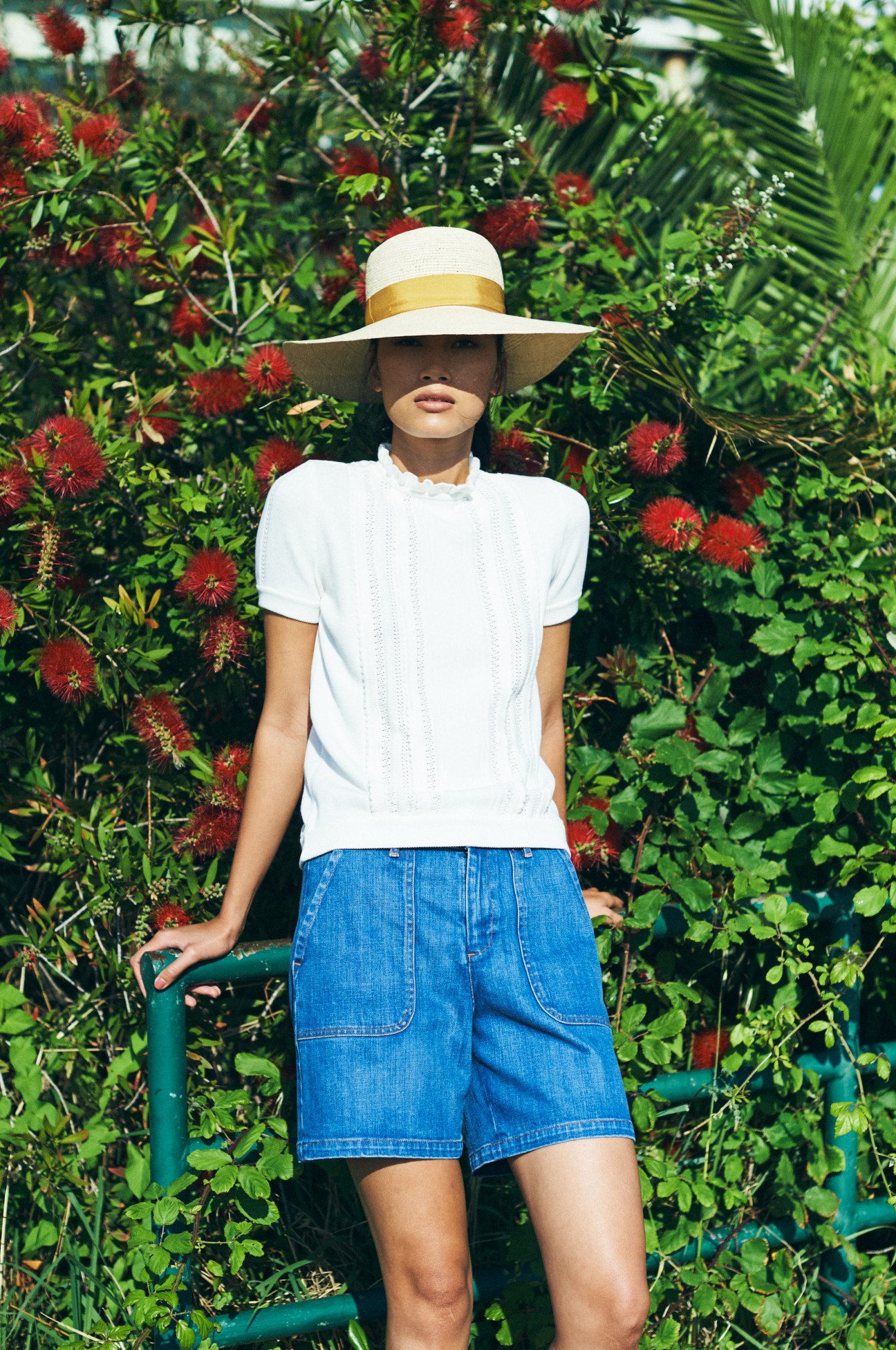 Woman wearing white top and blue denim shorts with white wide-brimmed hat against red flowering plants.