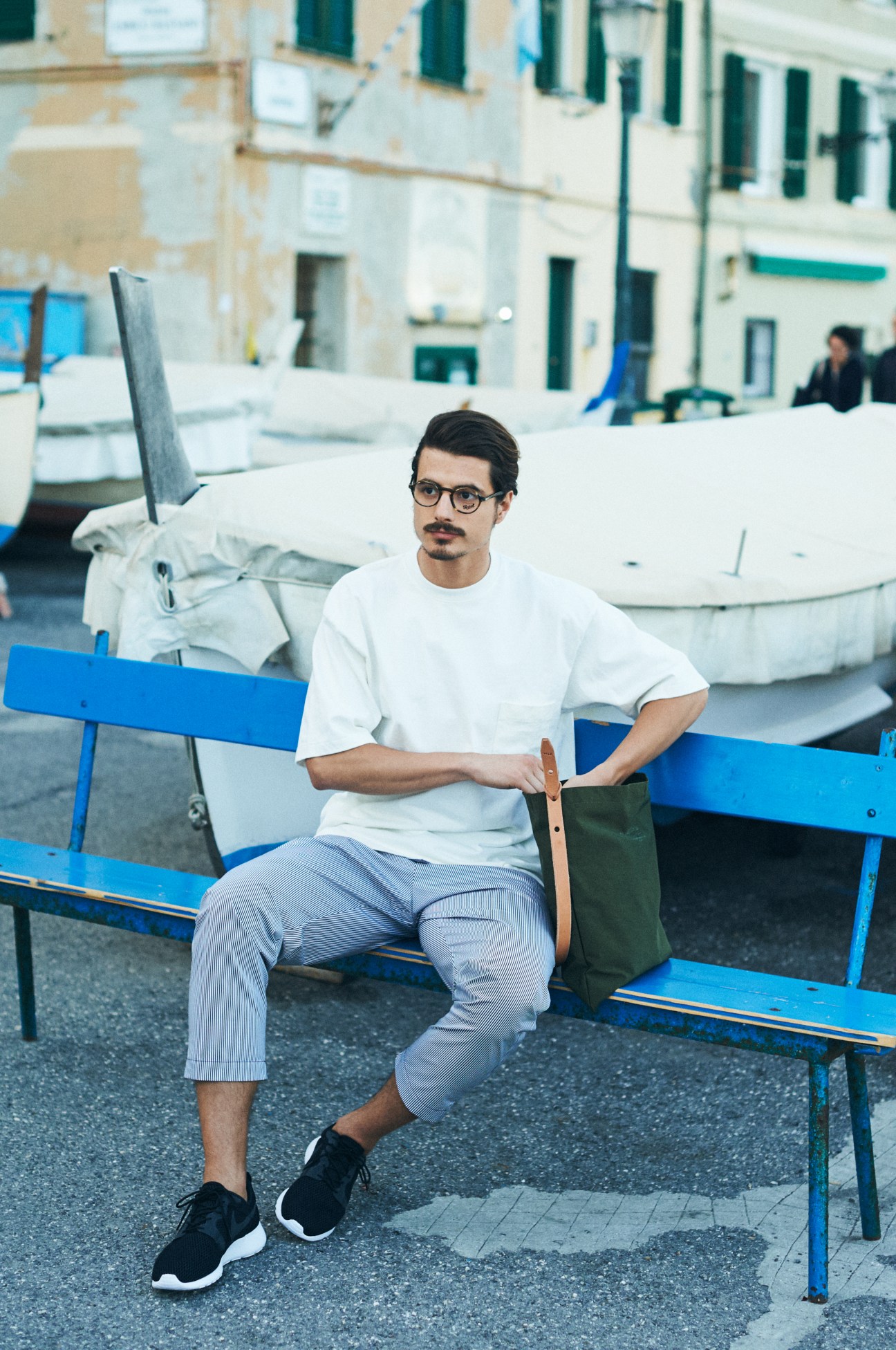 Man in white t-shirt and light blue trousers sitting on a blue bench by Genoa's waterfront with boats in background.