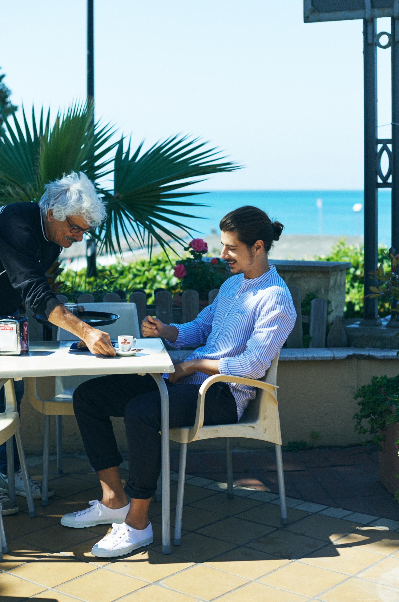 Two men seated at a waterfront table overlooking the sea in Genoa, one wearing an Oliver Spencer shirt and Ermenegildo Zegna trousers.