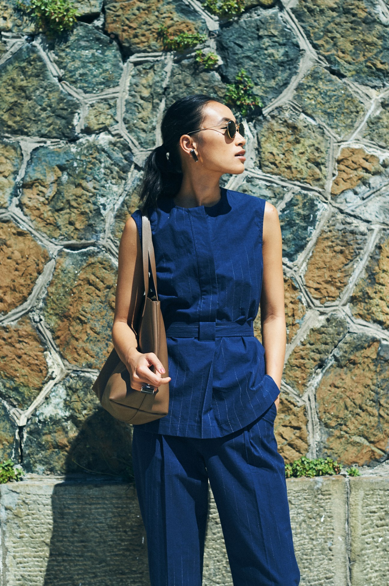 Woman in navy pinstriped sleeveless top and trousers standing against a stone wall with ivy.