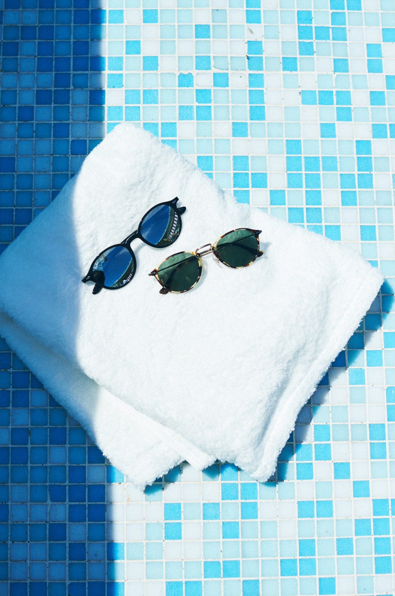 Two pairs of sunglasses resting on a white towel by a blue and white tiled pool.