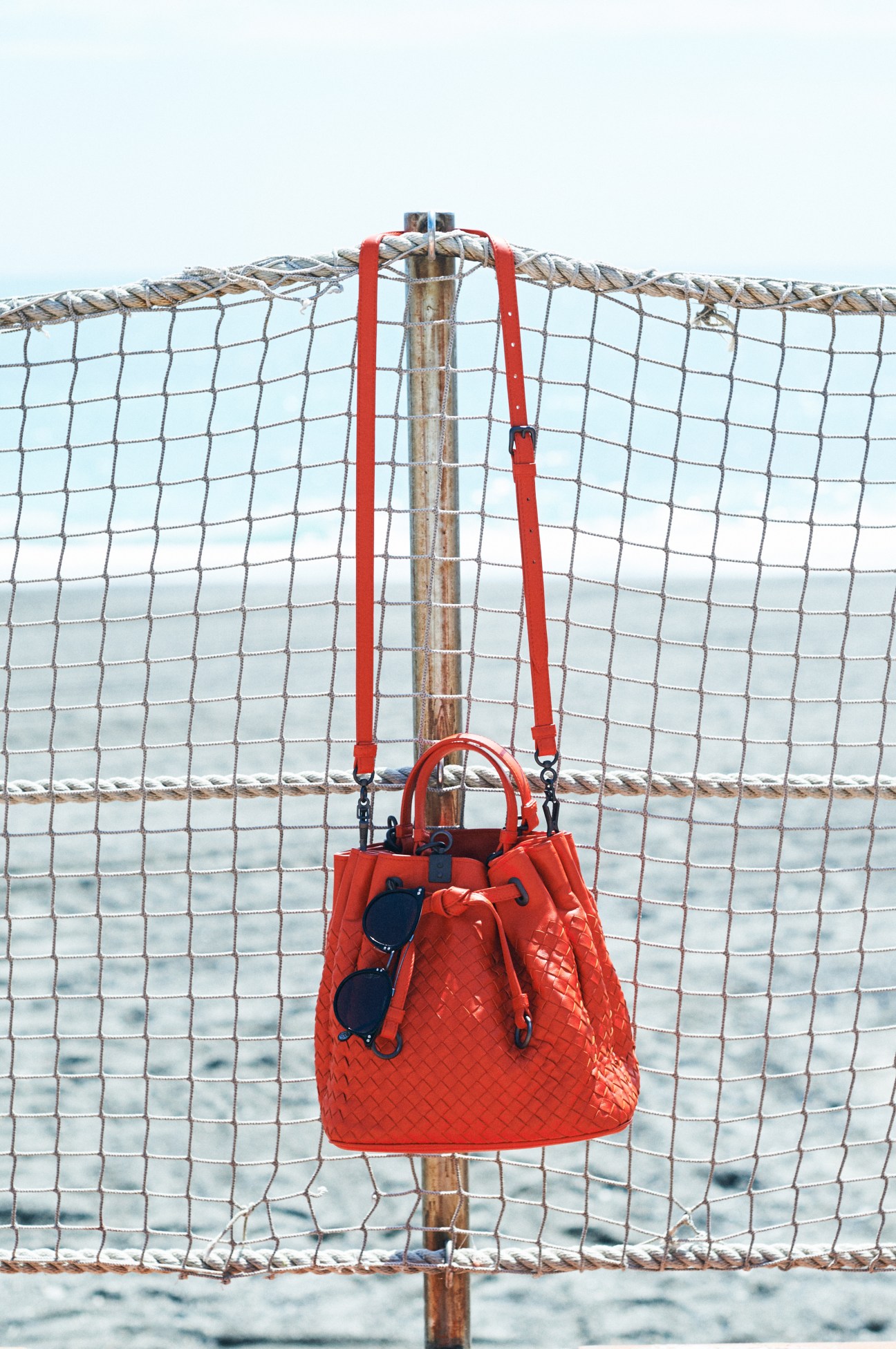 Red woven bag with sunglasses hanging from rope against beach volleyball net and sea.