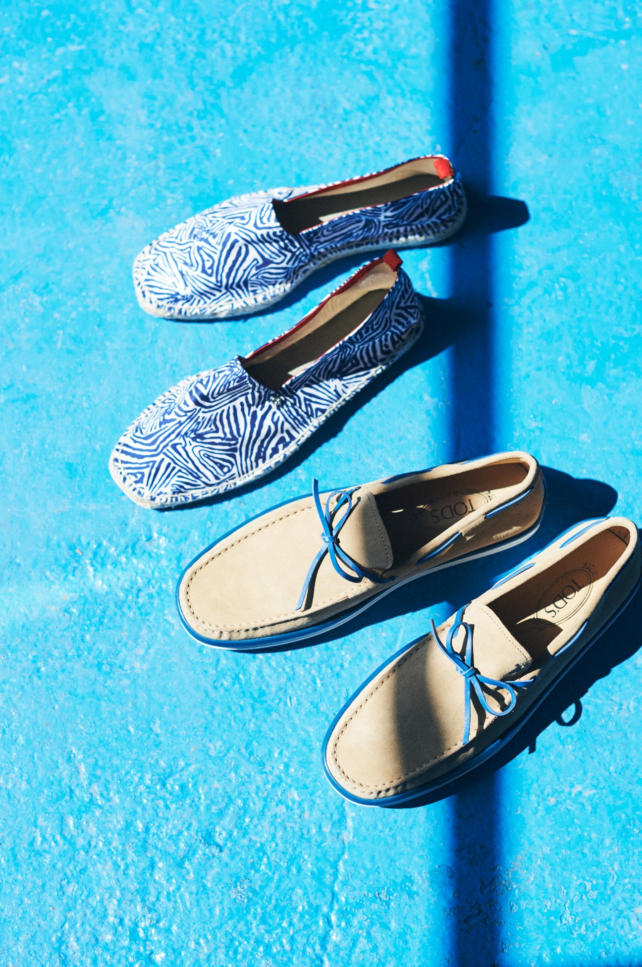 Two pairs of slip-on shoes displayed on a bright blue background: navy and white patterned slip-ons by Orlebar Brown on top, cream leather boat shoes by Tod's below.