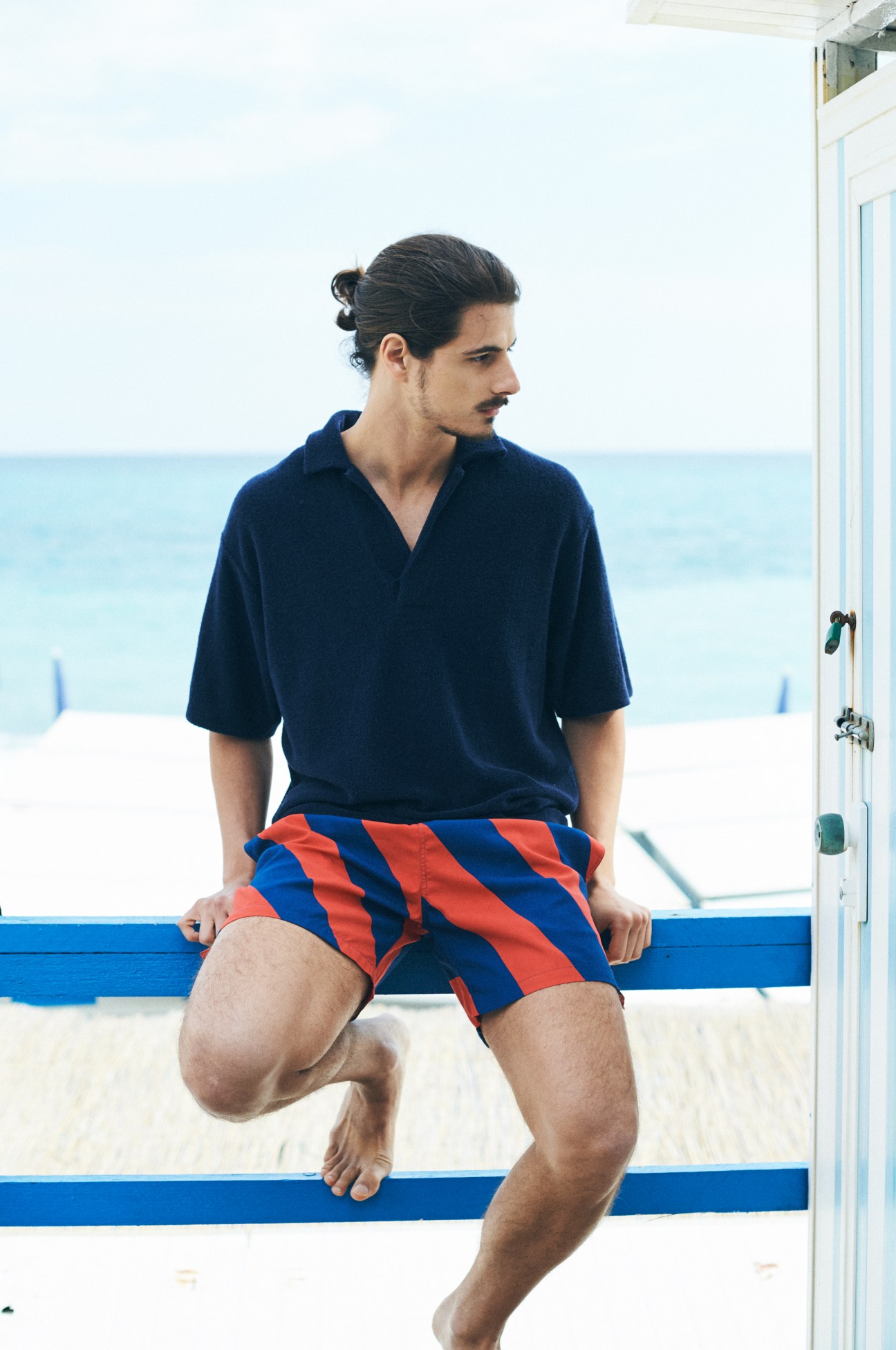 Man in navy polo shirt and red-and-blue striped swim shorts sitting on blue beach railing overlooking the sea.