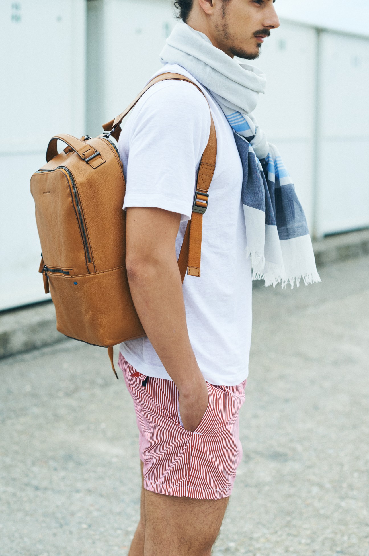Man wearing tan leather backpack, white t-shirt, red striped shorts, and patterned scarf, shown from side profile.