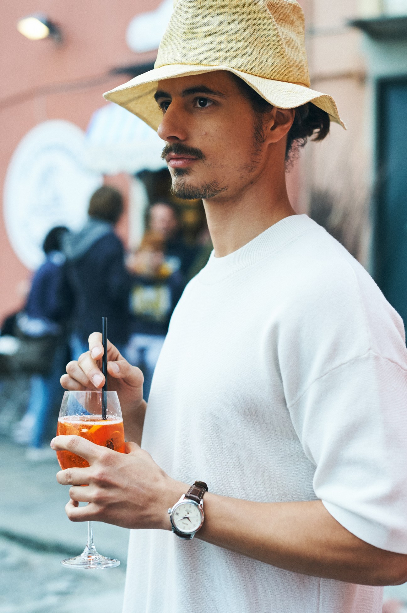Man wearing a beige hat and white t-shirt holding a cocktail on a Genoa street.
