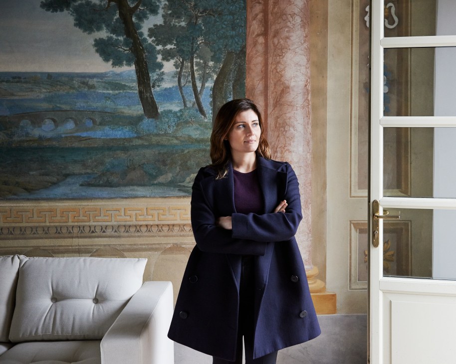 Gaia Gaja stands in an elegant palazzo interior with arms crossed, wearing a dark blazer.