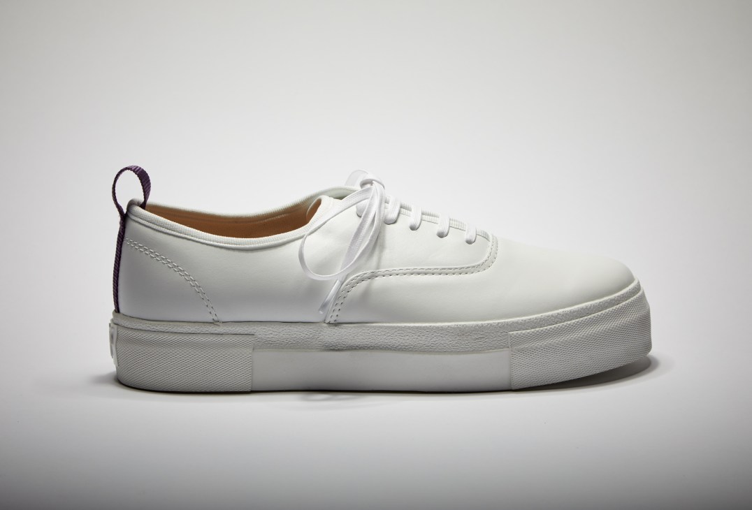 White leather sneaker with platform rubber sole and purple heel tab, shown in profile against neutral background.