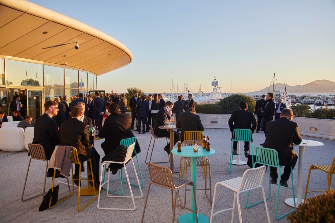 Guests in business attire mingle on a rooftop terrace with colorful modern furniture overlooking a harbor and yachts in Cannes.