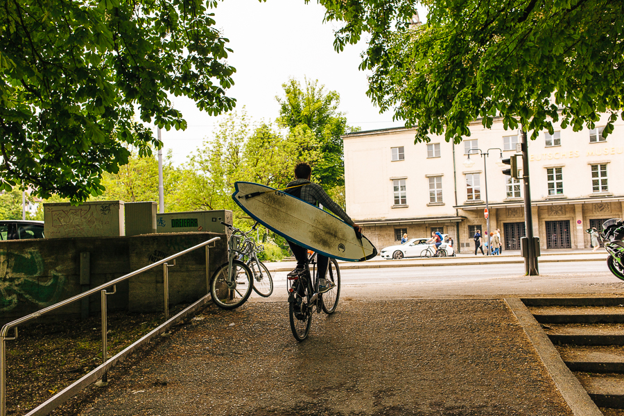 Cyclist carrying a surfboard walks bike through urban plaza with trees and historic buildings
