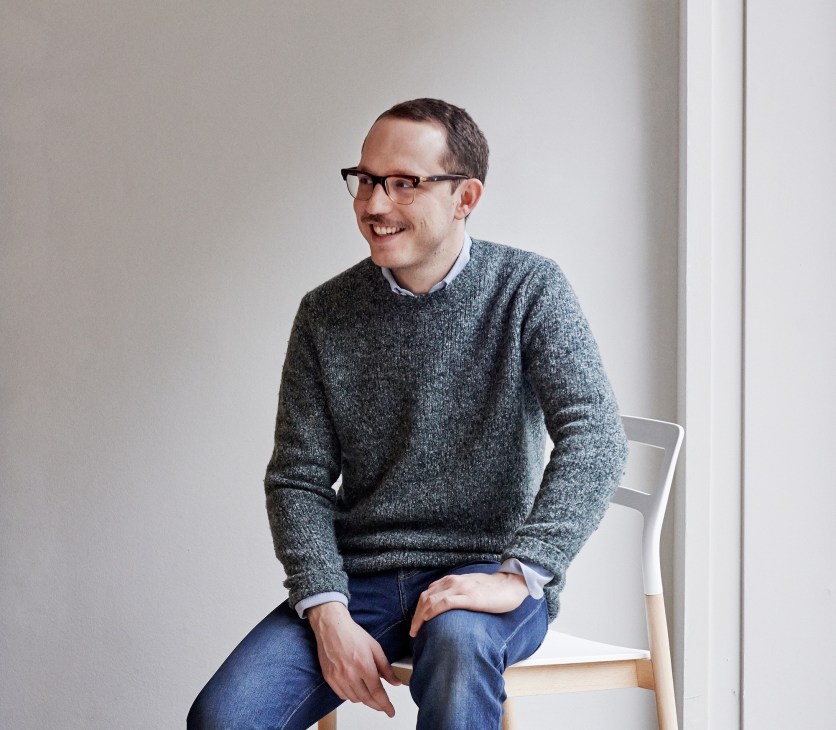 Marco Velardi, creative director of De Padova, seated on a white chair wearing glasses and a grey sweater.