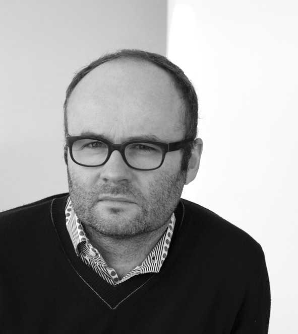 Black and white portrait of a man wearing glasses and a dark sweater over a striped shirt.