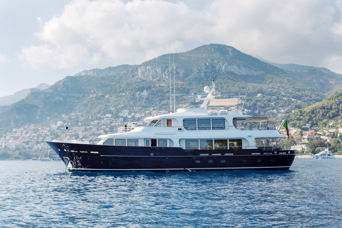 White and dark blue luxury yacht anchored in Mediterranean waters with mountainous coastline in background.