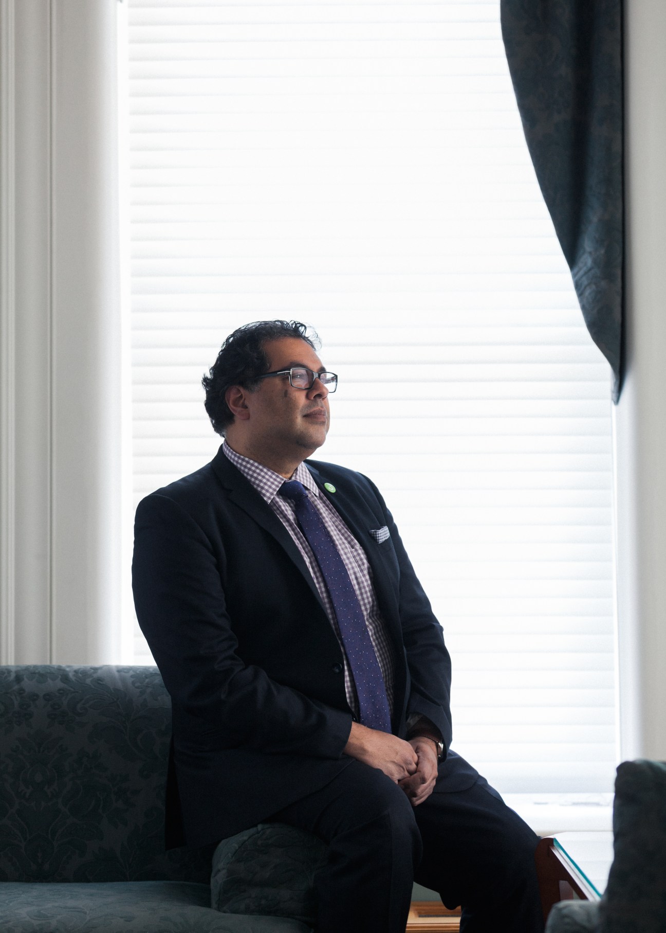 Naheed Nenshi, Mayor of Calgary, sits by a window in his office wearing a navy blazer and checkered shirt.