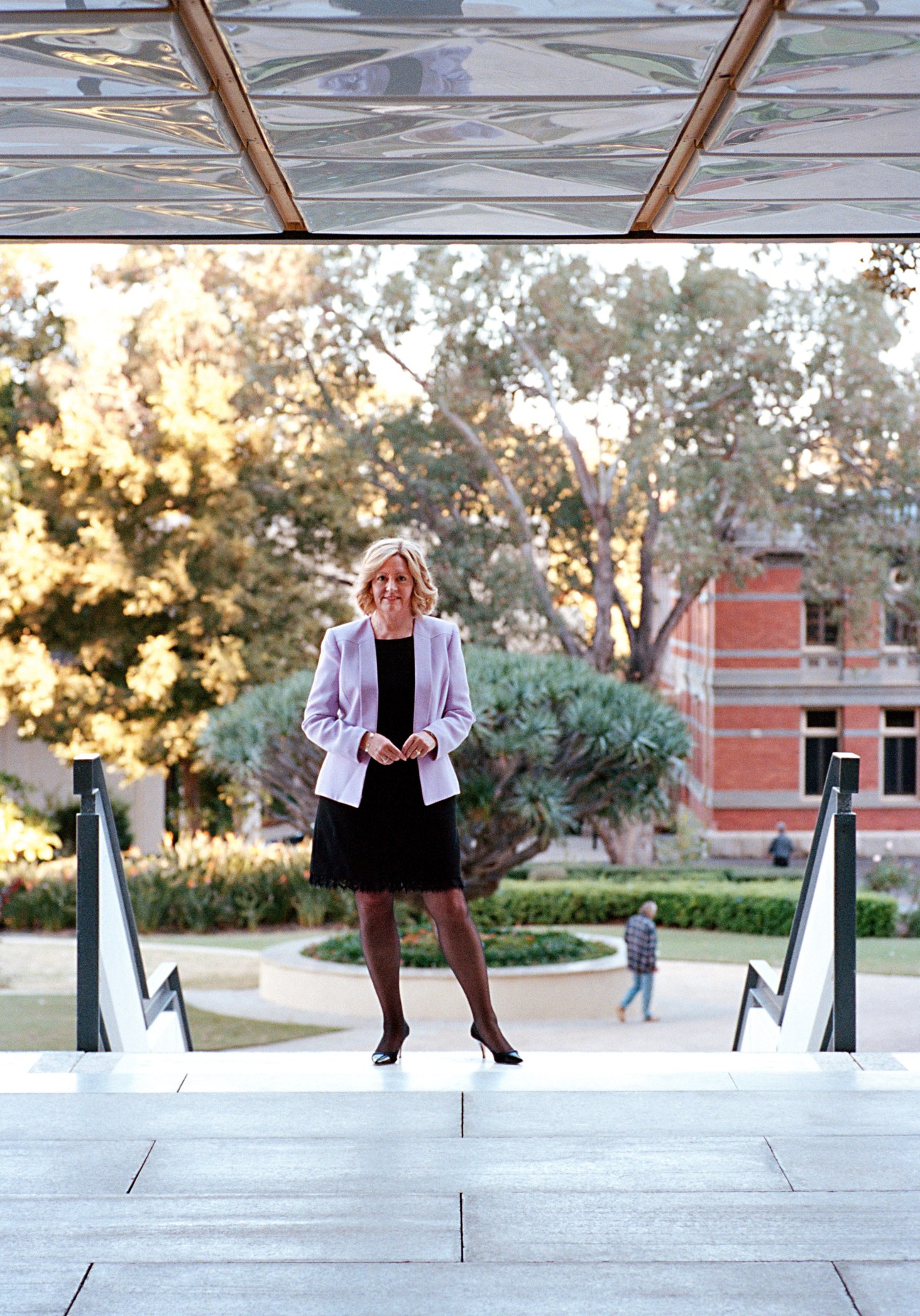 Lisa Scaffidi, Lord Mayor of Perth, stands on steps beneath a glass canopy in a public plaza with trees and a brick building behind her.