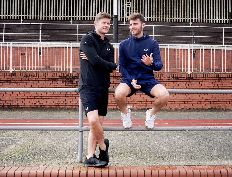 Two men in Castore athletic wear exercising outdoors against a brick wall and metal fence.