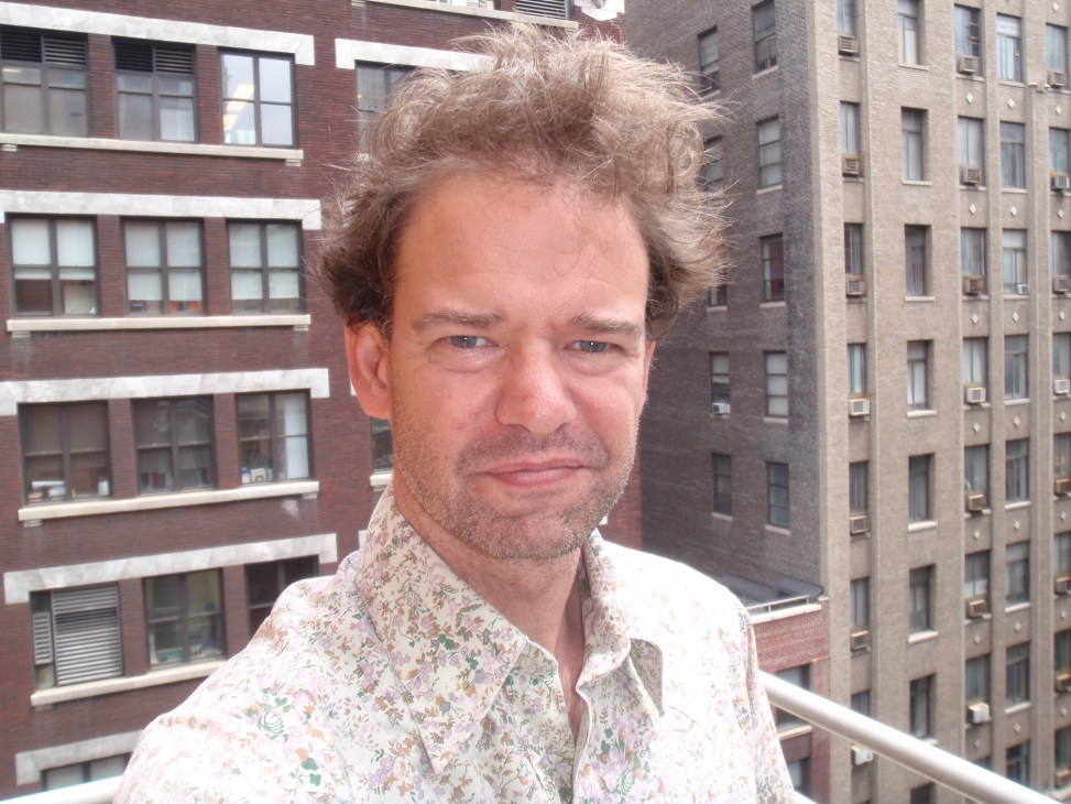 Man in patterned shirt on balcony overlooking brick buildings in urban setting.