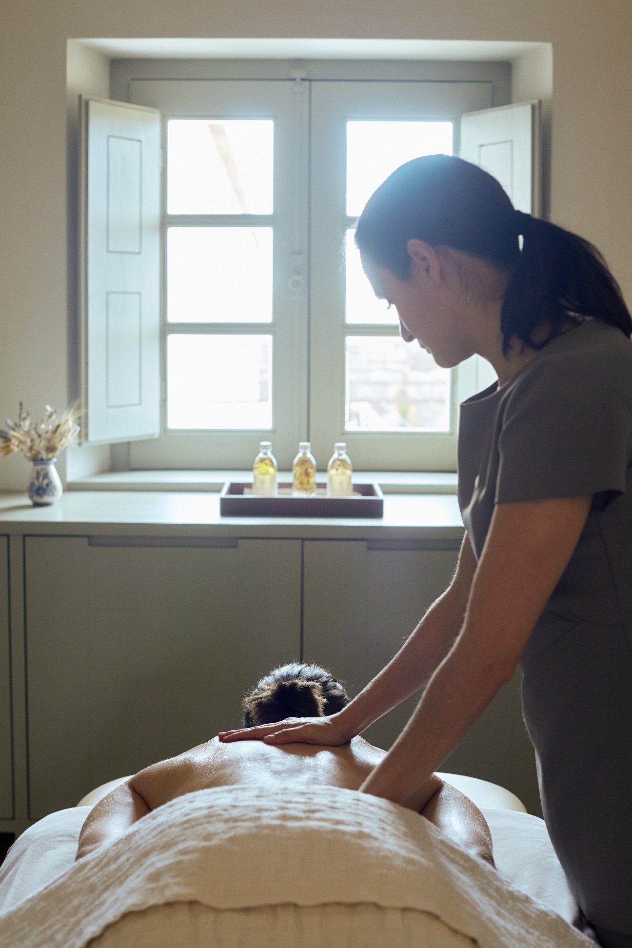 A spa therapist performing a massage on a client in a bright, minimalist room with whitewashed walls and large windows.