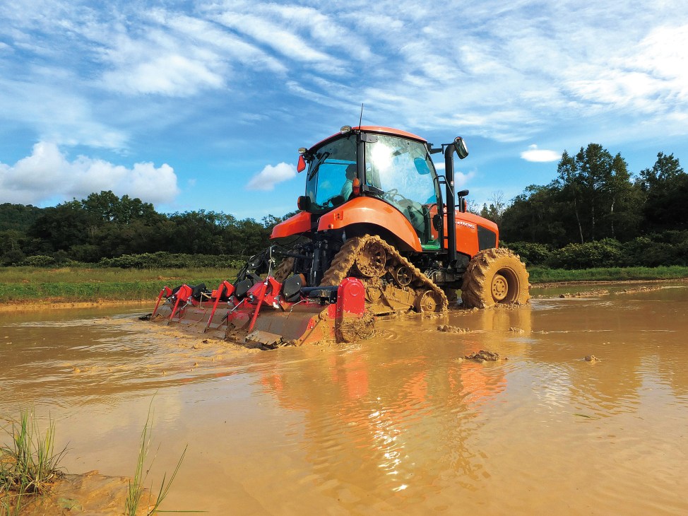Orange and red Kubota tractor with red planting equipment in muddy water during field operations.
