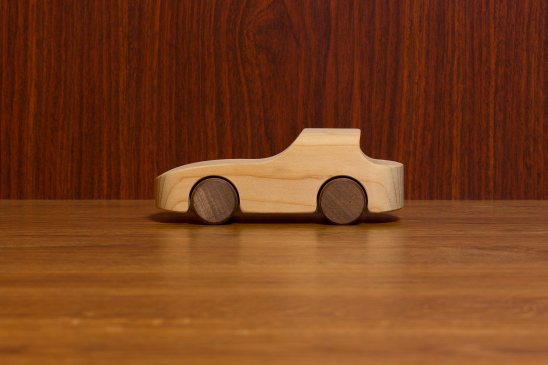 Hand-crafted wooden toy car in natural light wood with darker wooden wheels against a wood grain background.