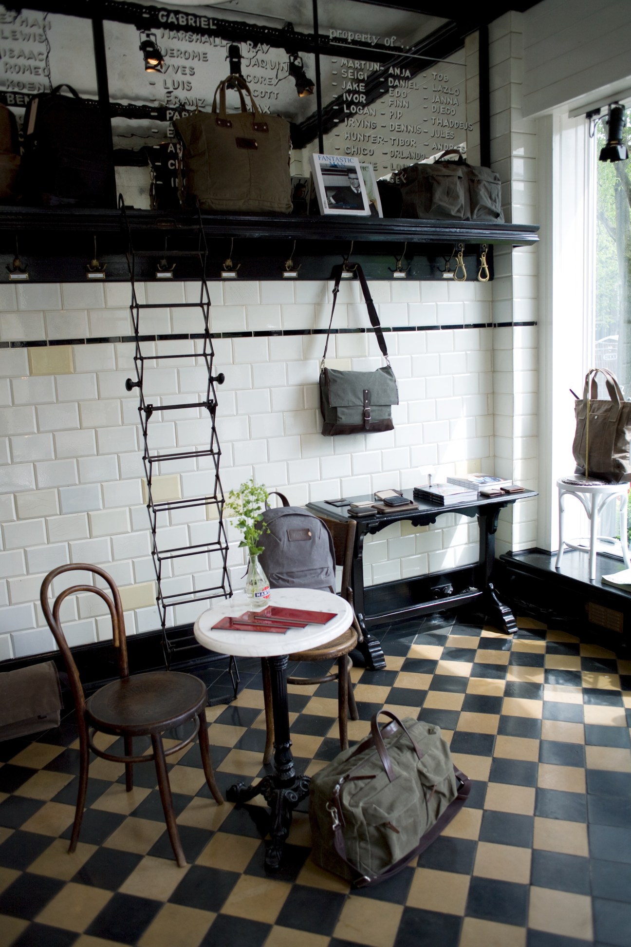 Interior of Property Of… store in Amsterdam showing white subway tile walls, black shelving with men's bags, and checkered floor with furniture.