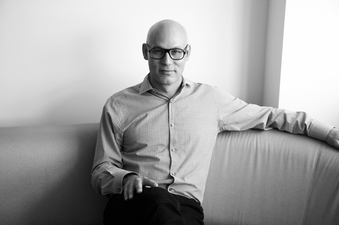 Craig Robins, a bald man wearing glasses and a checkered shirt, seated on a sofa.