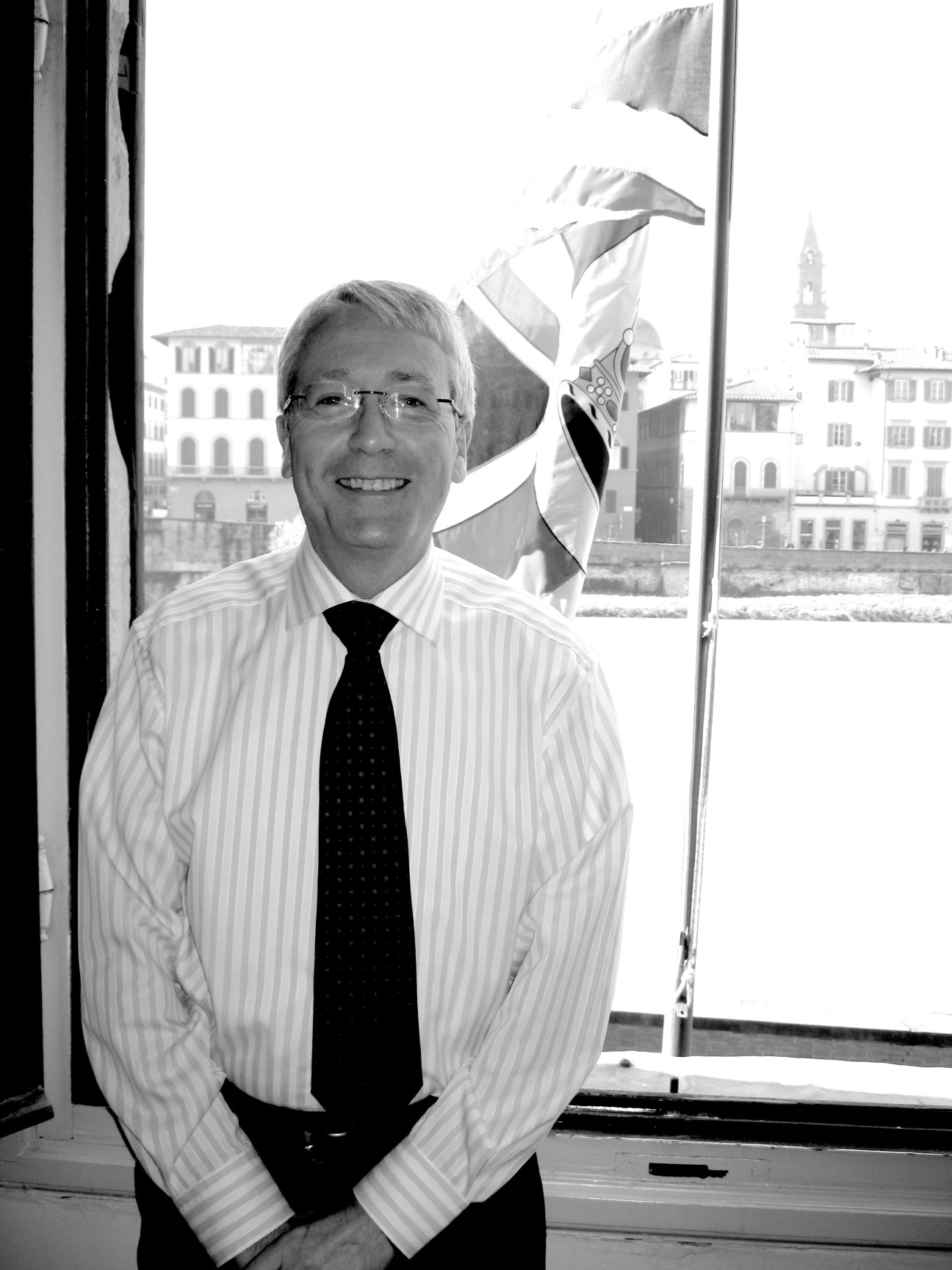 Portrait of David Broomfield, British consul in Florence, standing by a window with Italian cityscape and flags visible behind him.