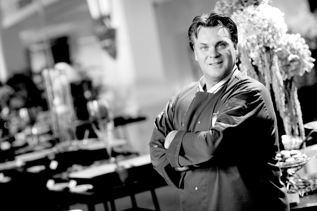 François Payard, owner of Payard pastry shop, poses in a formal black and white portrait at his kitchen counter surrounded by white flowers and chocolate confections.