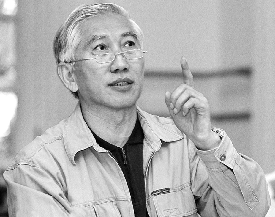 Asian man wearing glasses and light jacket gestures while speaking.