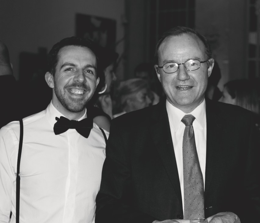 Two men in formal attire at an event, one wearing a tuxedo with bow tie and the other in a suit with tie.