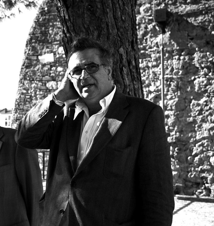 Man in sunglasses and dark blazer standing against ivy-covered stone wall, hand to face.