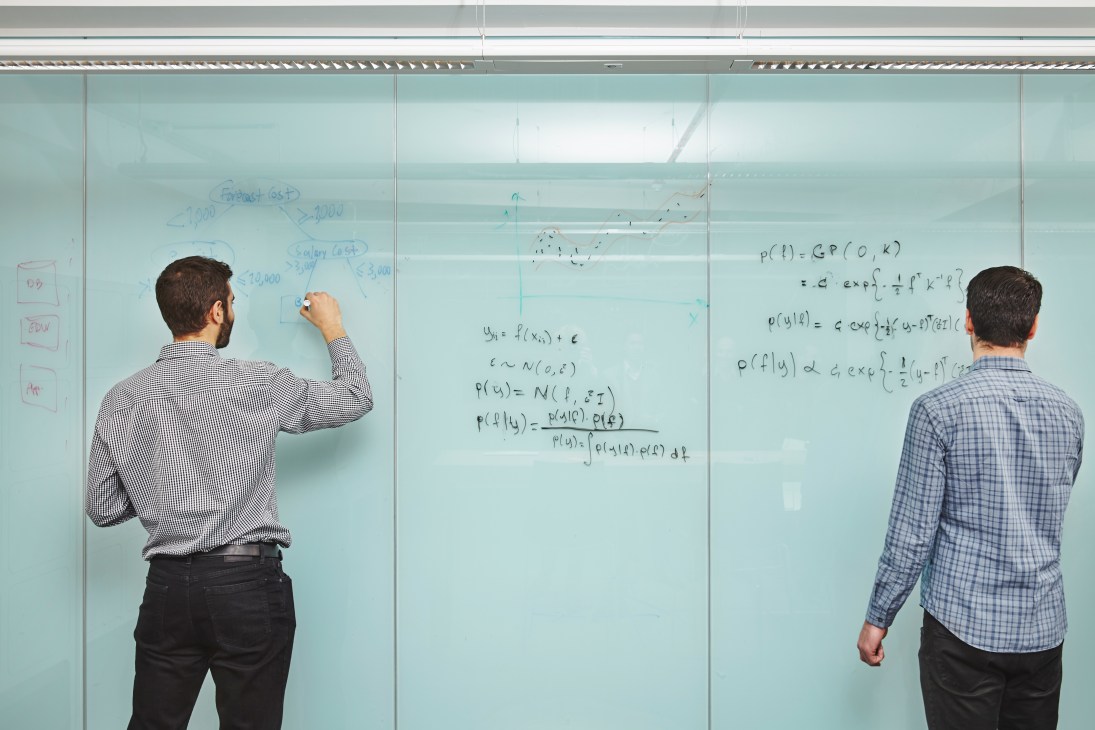 Two men in business casual attire writing mathematical equations and data analytics formulas on a glass whiteboard.