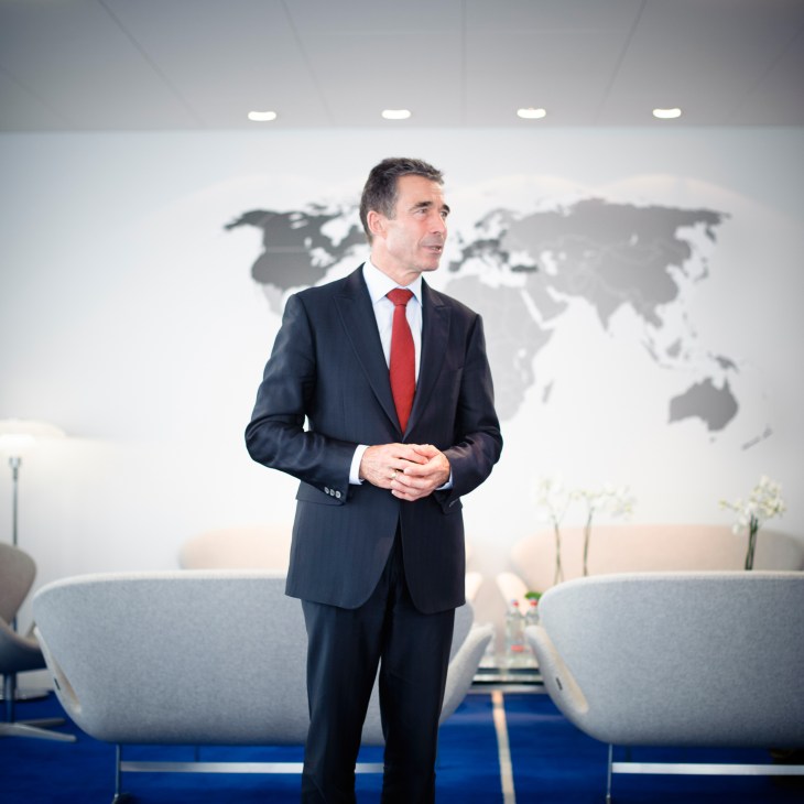 Man in dark suit with red tie standing in modern office with world map on wall behind him.