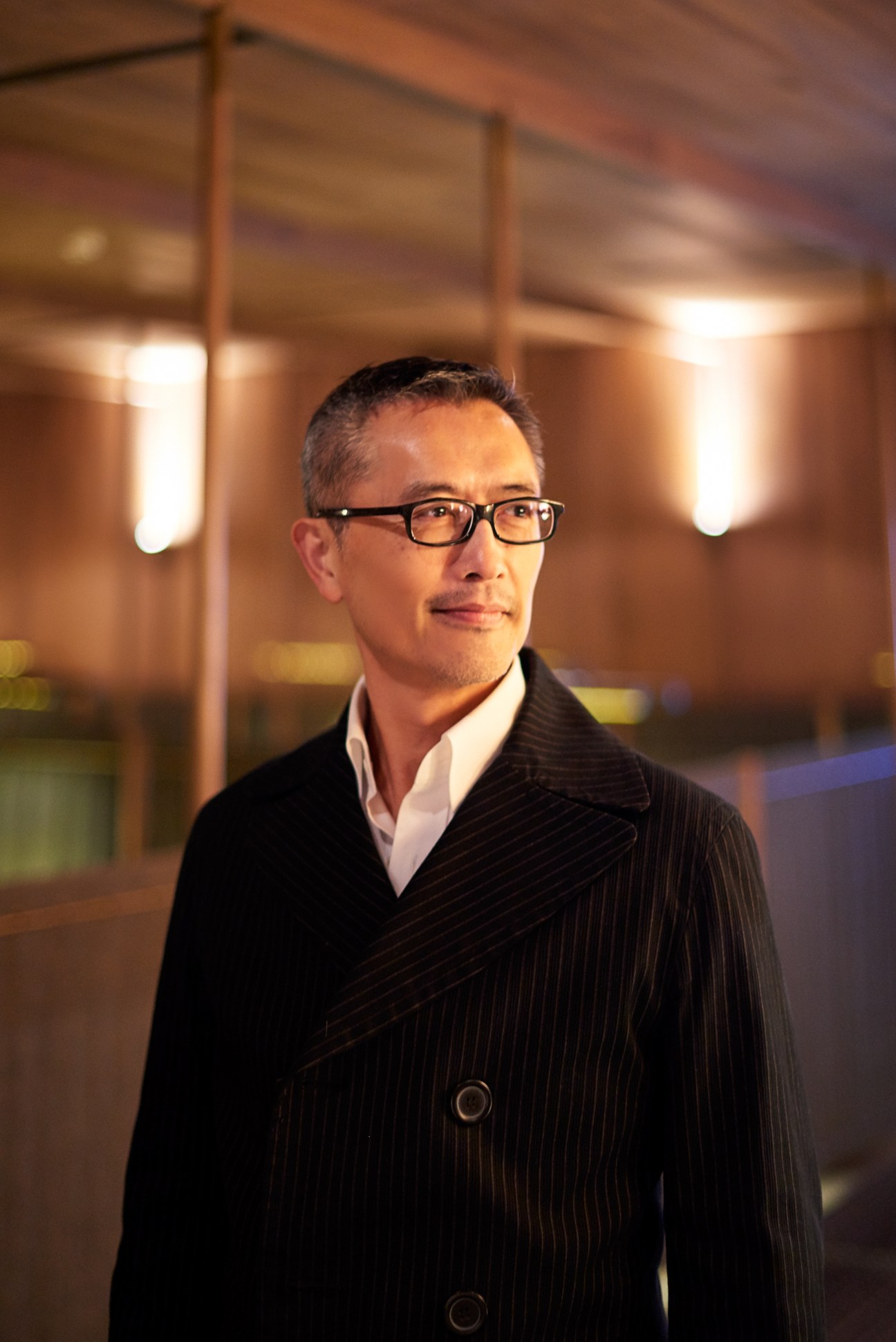 Portrait of Ray Chen, Taiwanese architect, wearing glasses and dark pinstriped jacket in modern interior space.