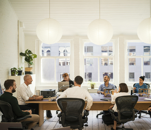 Team meeting at a wooden table in a bright office with high ceilings and large windows overlooking London.