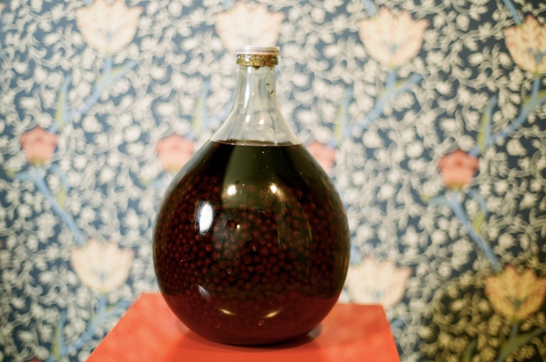 A round glass bottle filled with dark liquid against a patterned blue and white floral wallpaper background.