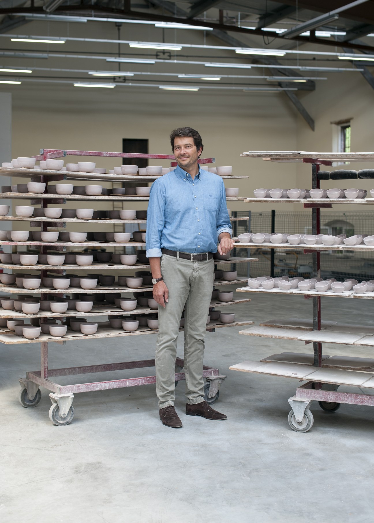 Olivier Passot stands in a porcelain factory surrounded by shelves of white bowls drying on racks.