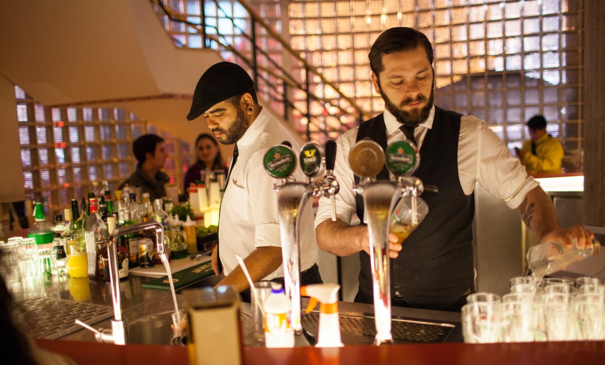 Two bartenders pour drinks at a counter in a modern venue with illuminated brick wall backdrop.