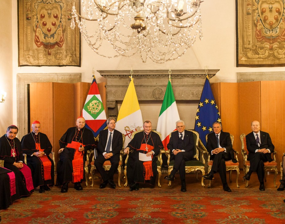 Eight men in formal black suits and robes seated beneath Italian, Vatican, and EU flags in an ornate ceremonial room.