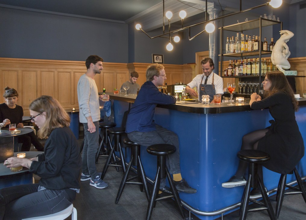 Interior of Central bar in Zürich with blue curved bar counter, pendant lighting, wood paneling, and customers and bartender at the counter.