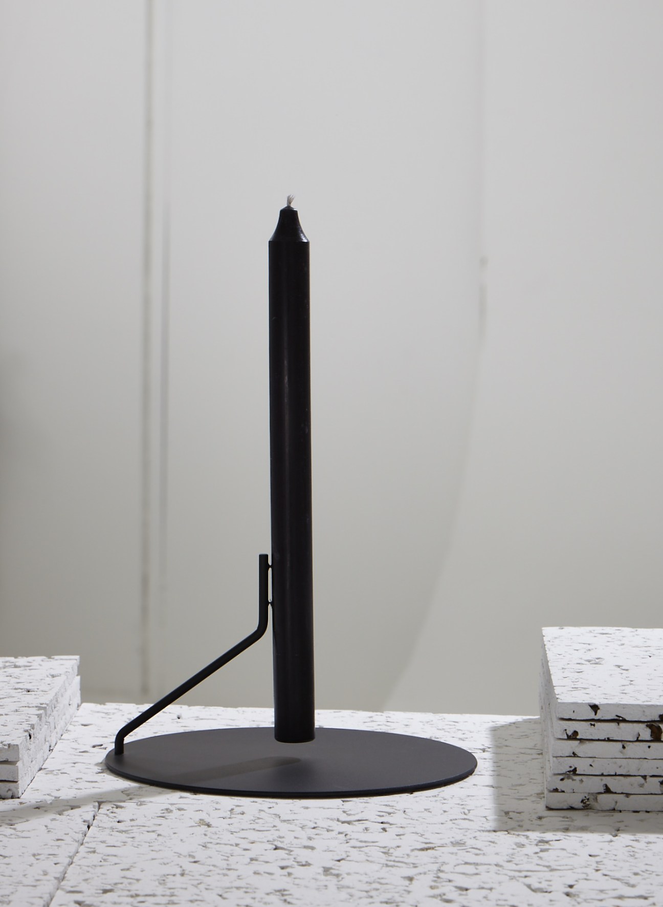 Black cylindrical candle in Buka candlestick with curved metal support on round base.