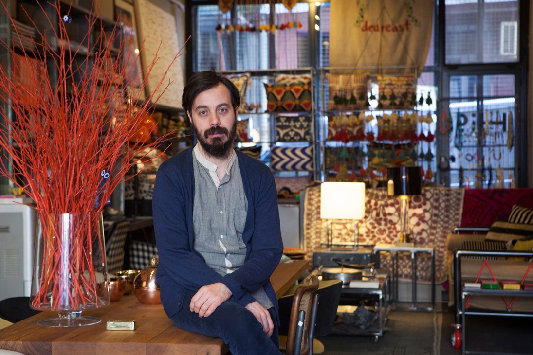 Man with dark beard sits in modern shop interior with red decorative branches and patterned textiles.