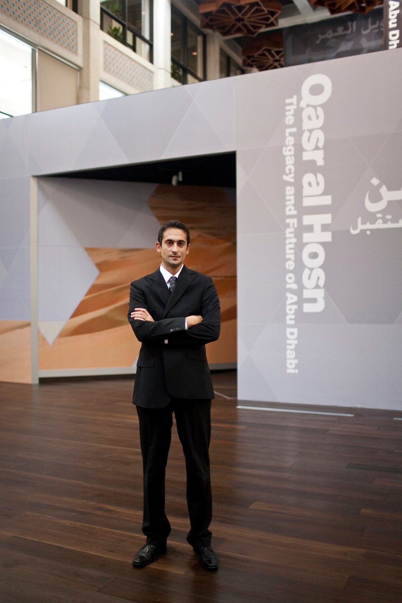 Man in black business suit standing with arms crossed in front of signage reading 'Saadiyat Host: The Legacy and Future of Abu Dhabi'
