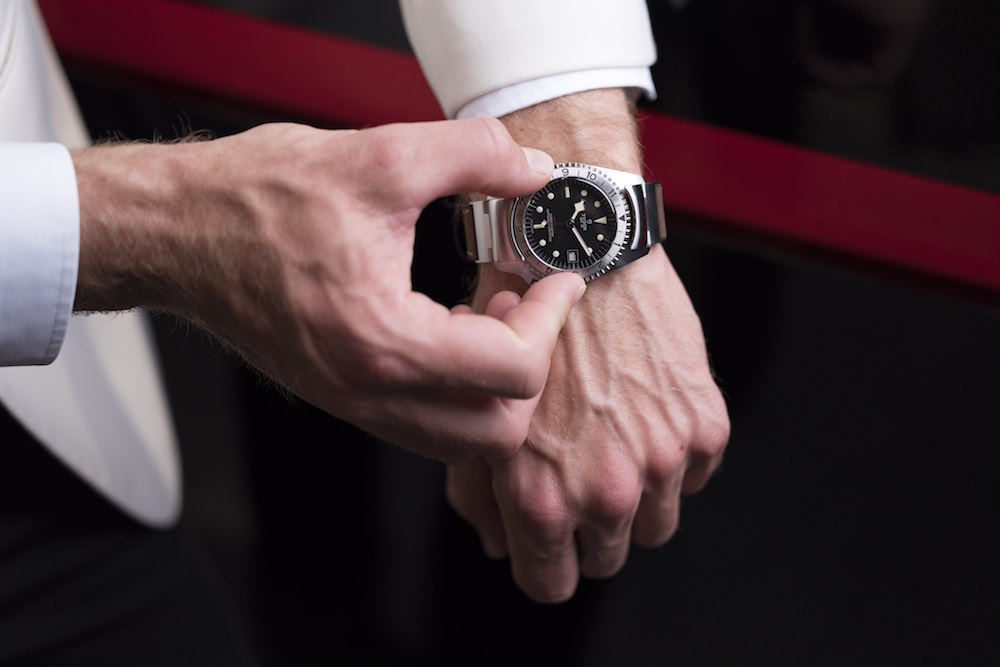 Man's hand wearing Tudor Black Bay P01 watch with black dial and hybrid leather-rubber strap against dark background.