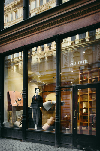 Samuji storefront with large windows displaying clothing on mannequins and merchandise, with black columns framing the entrance.