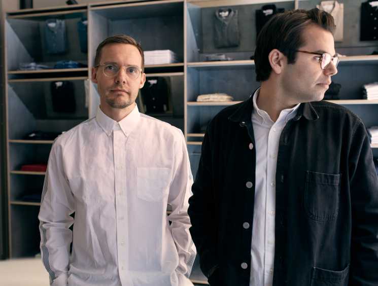Two men in a clothing boutique stand in front of wooden shelves stocked with folded shirts. One wears a white button-down with striped cuffs; the other wears a white shirt under a dark denim shacket and glasses, looking off to the side.
