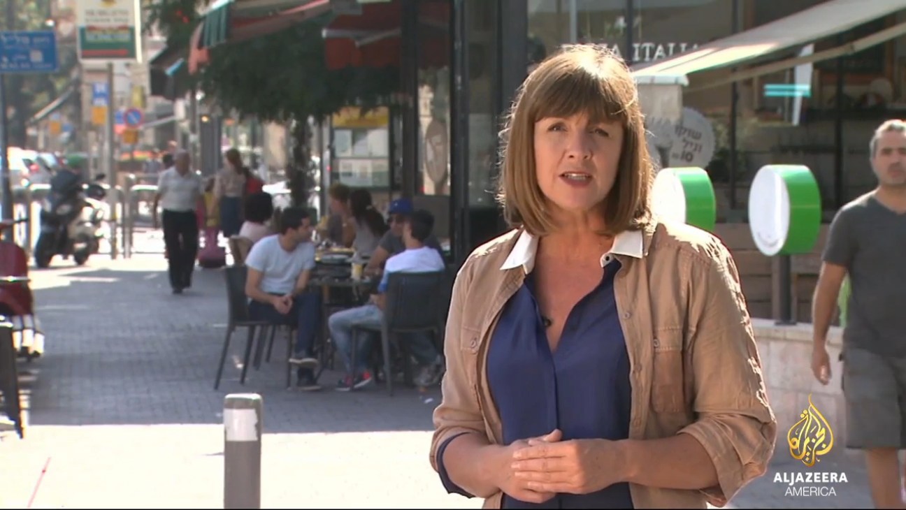 Al Jazeera America news presenter broadcasting from a street location with pedestrians and storefronts visible in the background.