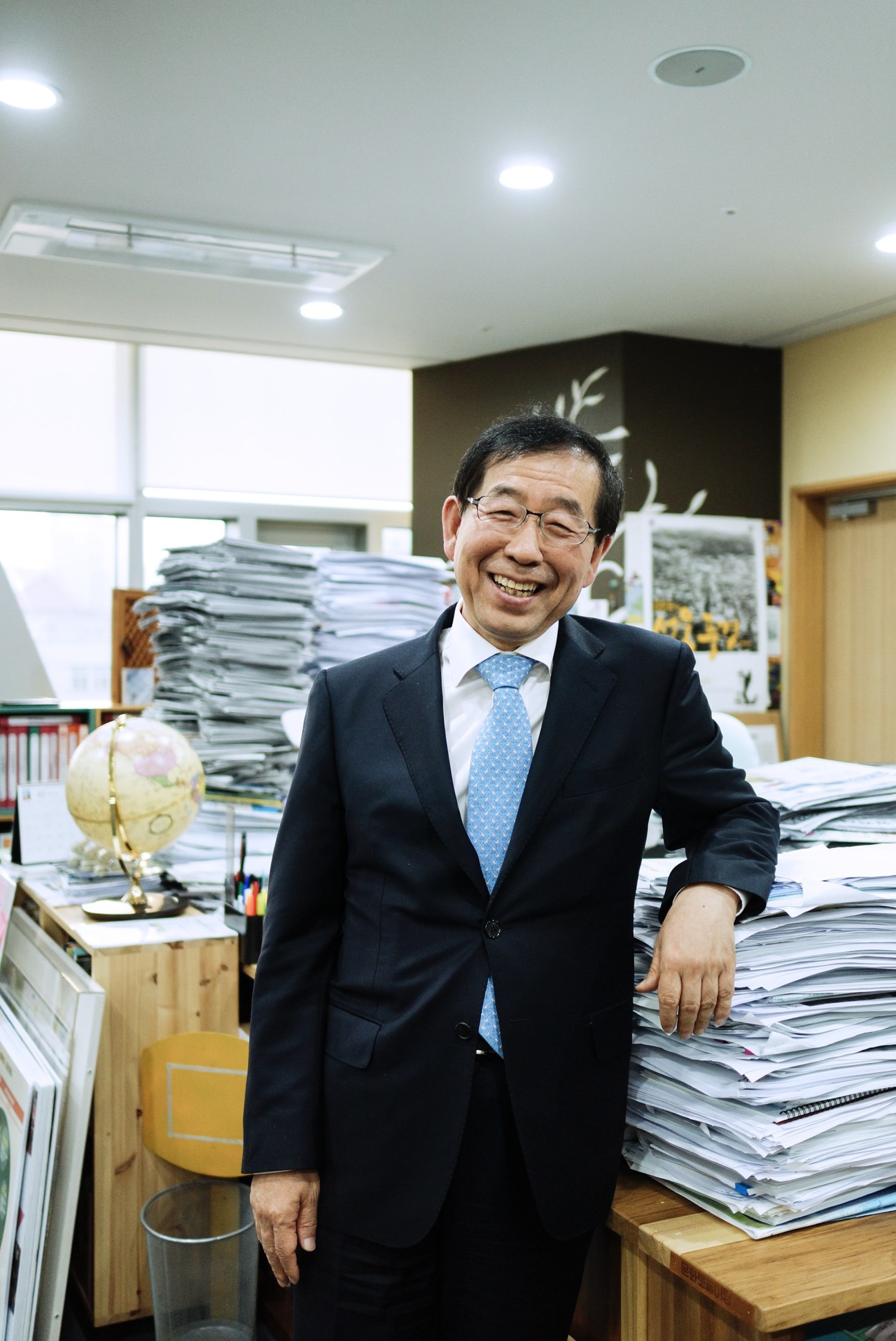 Park Won-Soon, mayor of Seoul, smiling in his office surrounded by stacked documents and papers.
