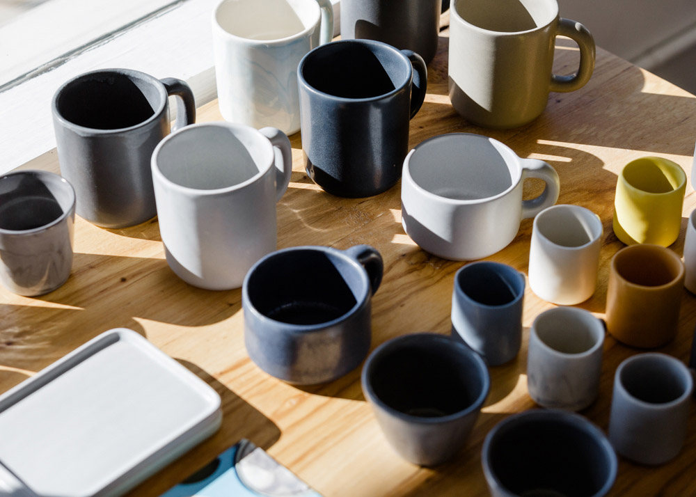 Assorted ceramic mugs in various colors and sizes displayed on a wooden table in natural light.