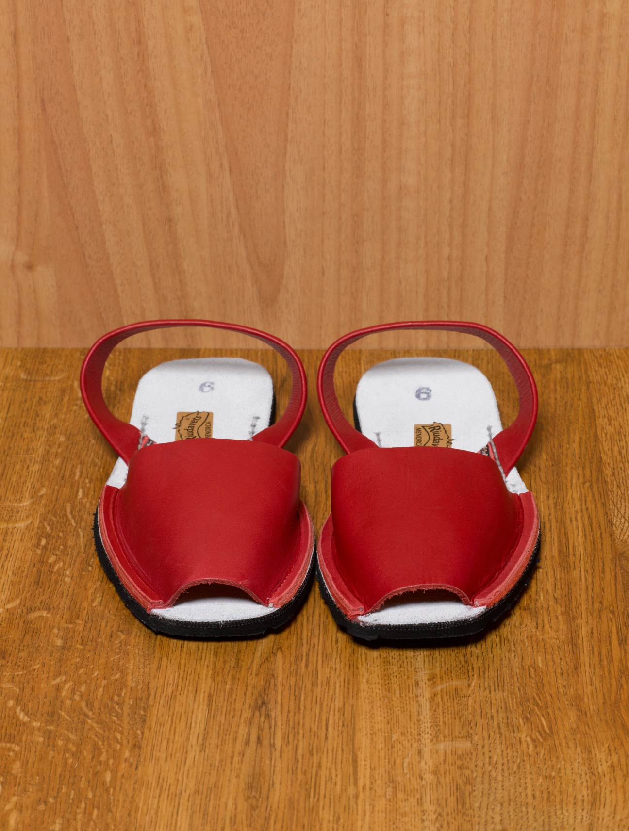 Pair of red Menorcan albarcas sandals with black soles on wooden floor.